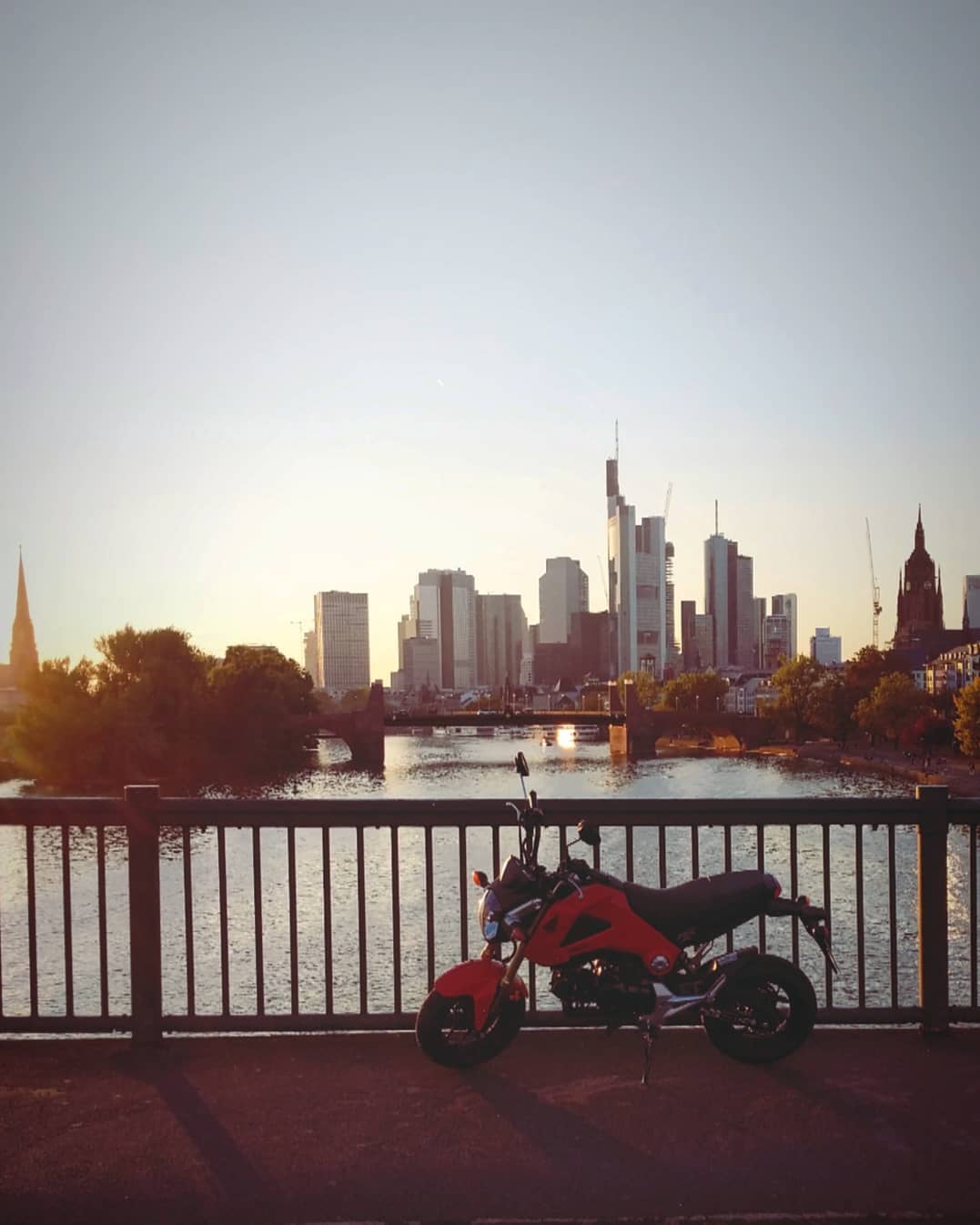 Frankfurt skyline at golden hour, with a little red Honda Grom parked on a bridge over the Main.